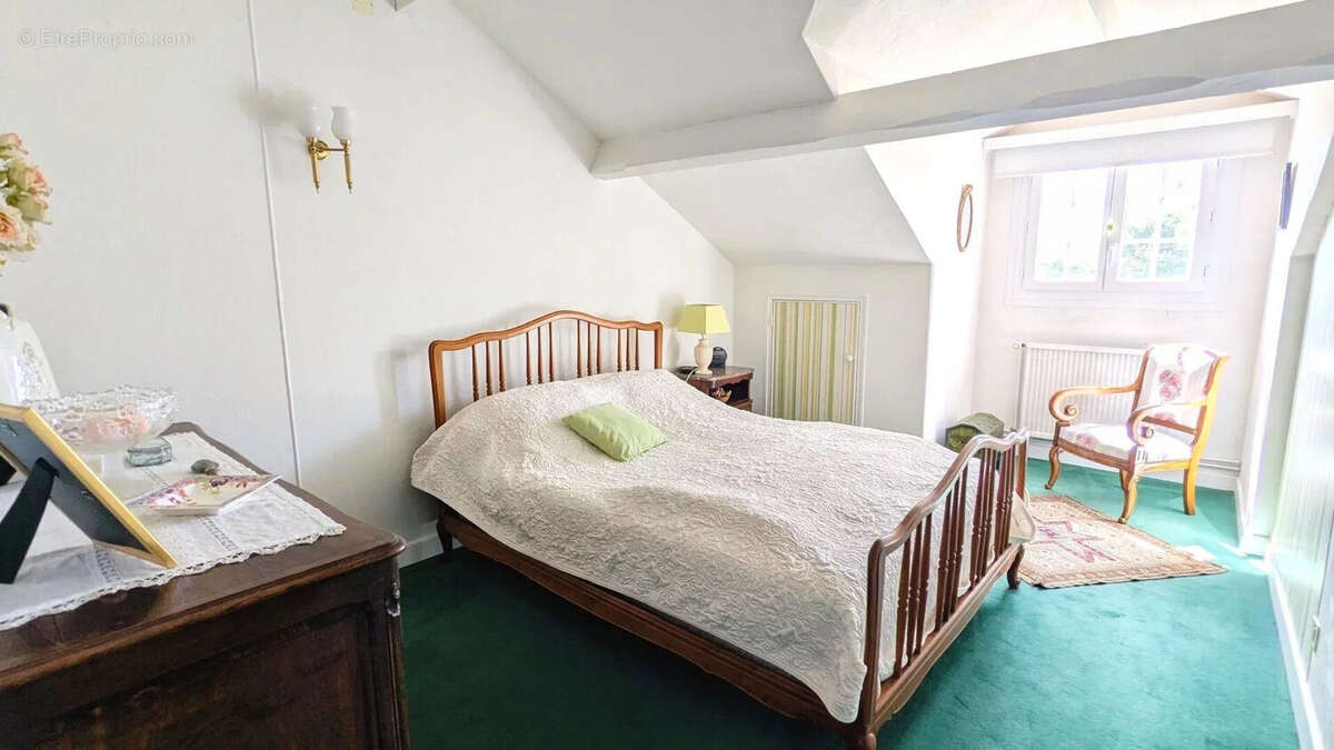Chambre lumineuse avec un lit double en bois, un fauteuil près de la fenêtre et un tapis vert sur le sol. - Maison à FOULAYRONNES