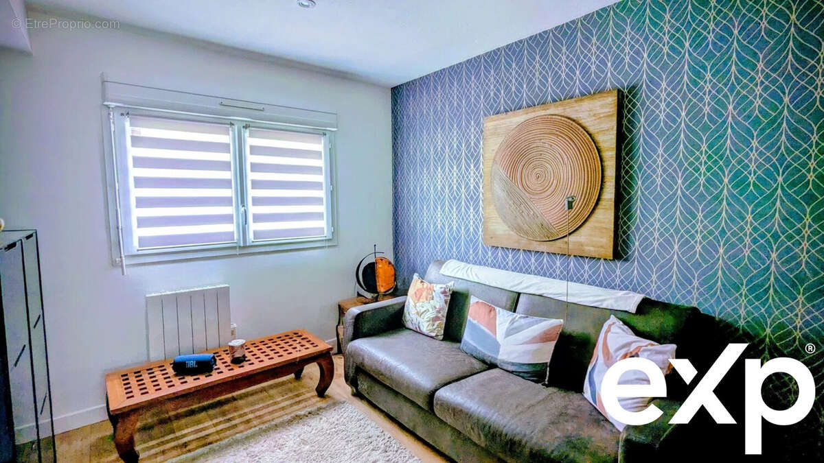 Un salon confortable avec un canapé gris, des coussins colorés, une table basse en bois et un tableau abstrait sur le mur bleu à motifs. - Maison à AIGUILLON