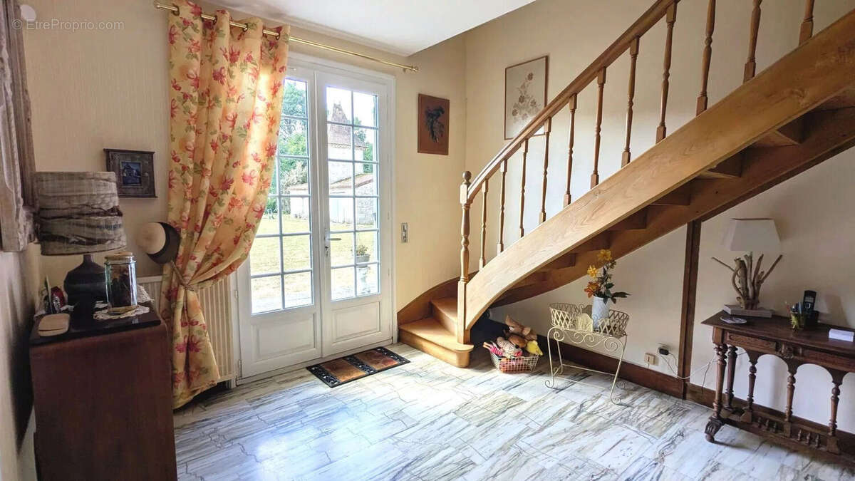 Une entrée lumineuse avec un escalier en bois et des portes-fenêtres donnant sur un jardin. Le sol est en carrelage clair et il y a quelques meubles et décorations. - Maison à FOULAYRONNES