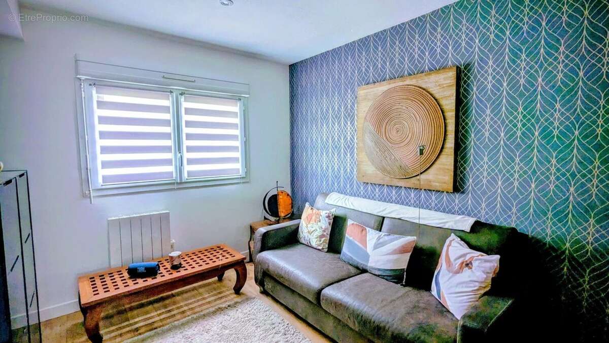 Un salon confortable avec un canapé gris, des coussins colorés, une table basse en bois et un tableau abstrait sur le mur bleu à motifs. - Maison à AIGUILLON