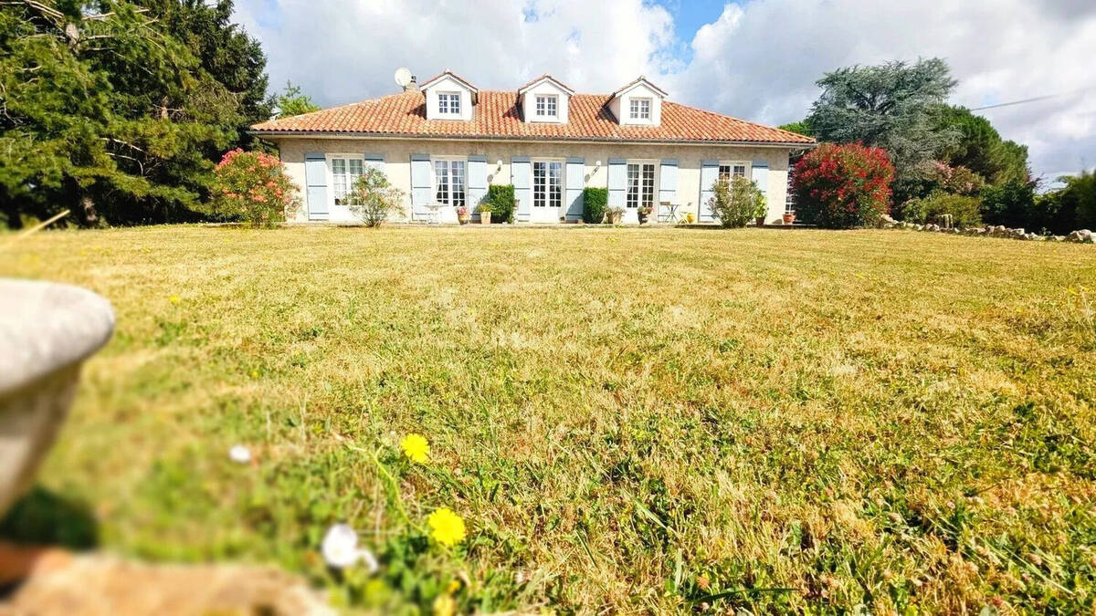 Une maison avec un toit en tuiles, entourée de verdure et de fleurs, sous un ciel partiellement nuageux. - Maison à FOULAYRONNES