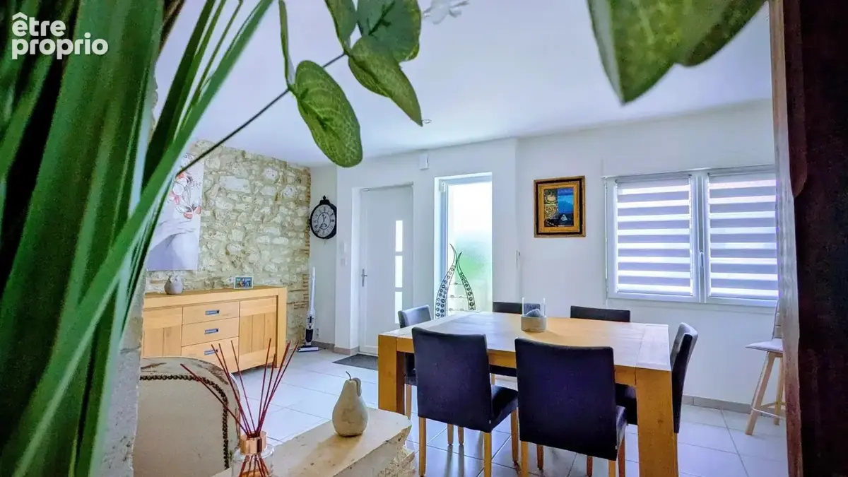 Salle à manger lumineuse avec table en bois, chaises noires, mur en pierre, et décorations végétales.