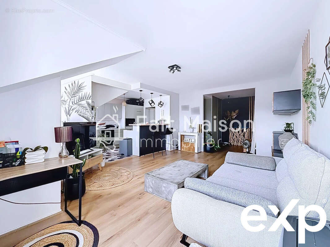 Un salon moderne et lumineux avec un canapé gris, une table basse, et un bureau sur la gauche. La pièce est ouverte sur une cuisine équipée. - Appartement à MASSY