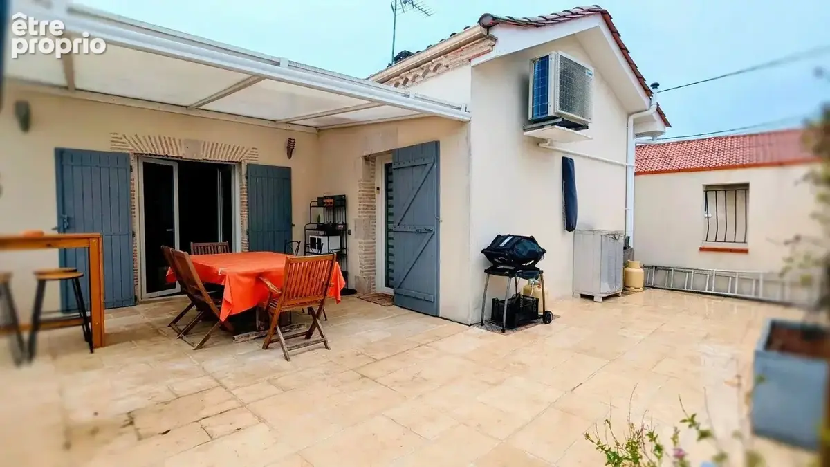 Une terrasse spacieuse avec une table à manger, des chaises en bois, un barbecue, et une table haute, devant une maison blanche avec des volets bleus. - Maison à AIGUILLON
