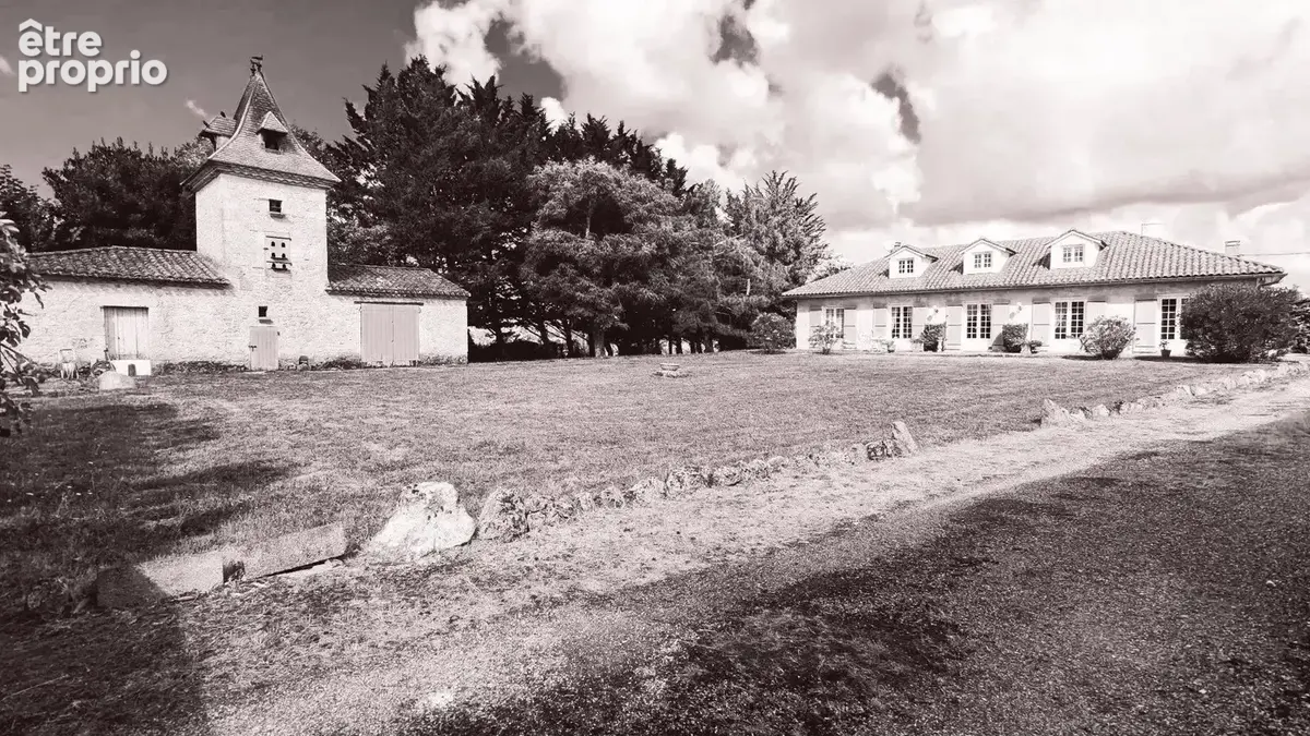 Une maison de campagne avec un bâtiment annexe en pierre, entourée d'arbres et sous un ciel partiellement nuageux.