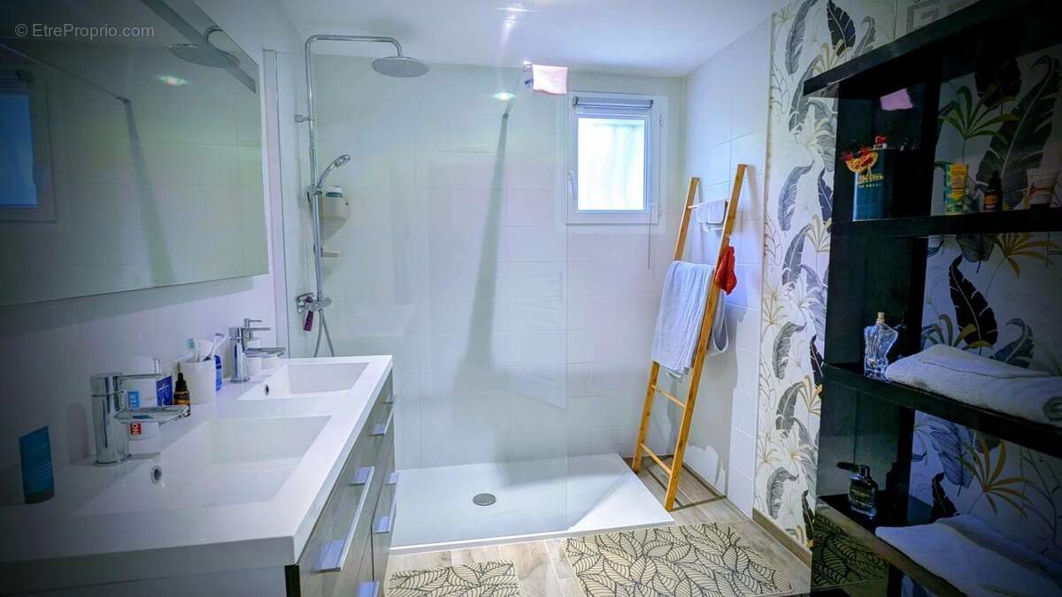 Une salle de bain moderne avec une douche spacieuse, un lavabo double, des étagères noires et des serviettes accrochées. - Maison à AIGUILLON