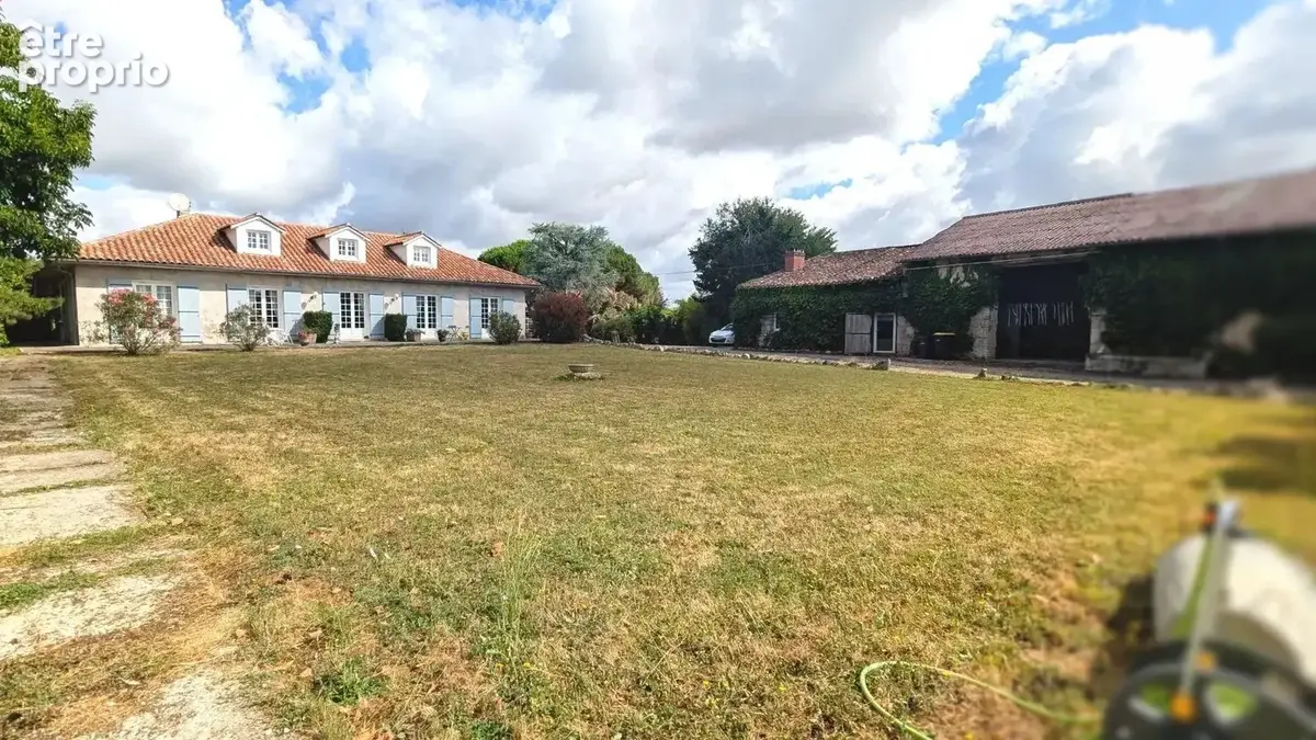 Une grande maison traditionnelle avec un toit en tuiles se trouve à gauche, à côté d'une grange recouverte de lierre à droite, entourée d'une vaste pelouse sous un ciel partiellement nuageux.