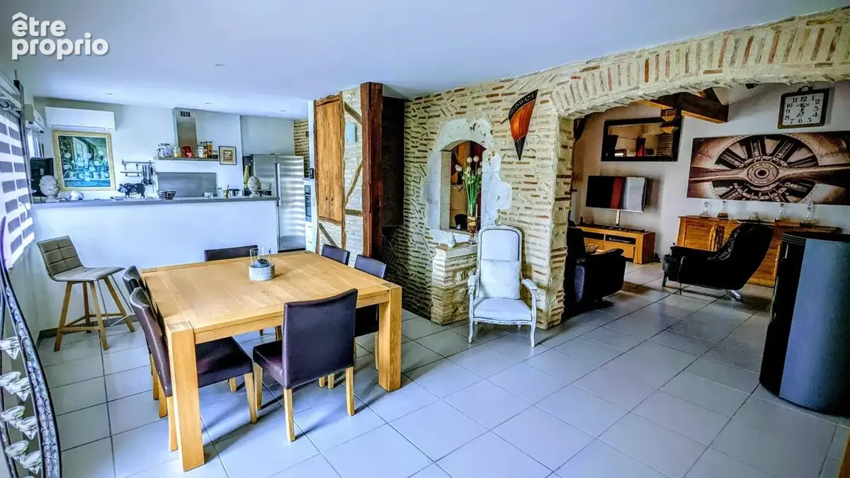 Une salle à manger ouverte avec une grande table en bois, des chaises noires, des murs en briques apparentes et une cuisine en arrière-plan. L'espace est moderne et lumineux.