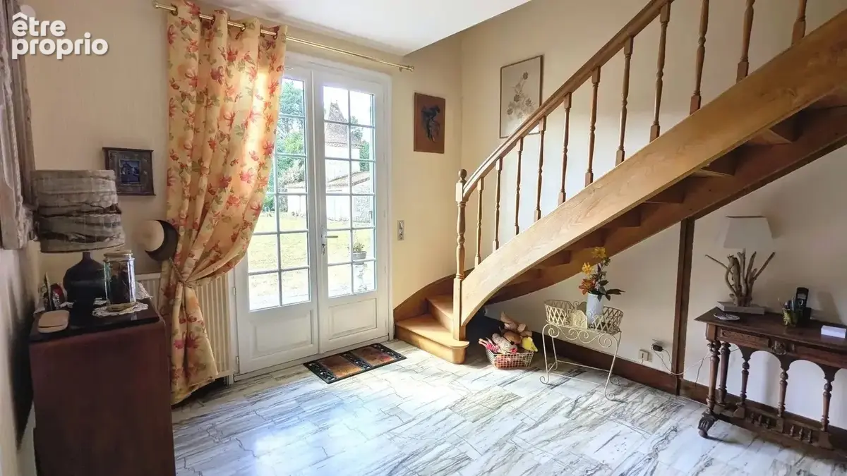 Une entrée lumineuse avec un escalier en bois et des portes-fenêtres donnant sur un jardin. Le sol est en carrelage clair et il y a quelques meubles et décorations.
