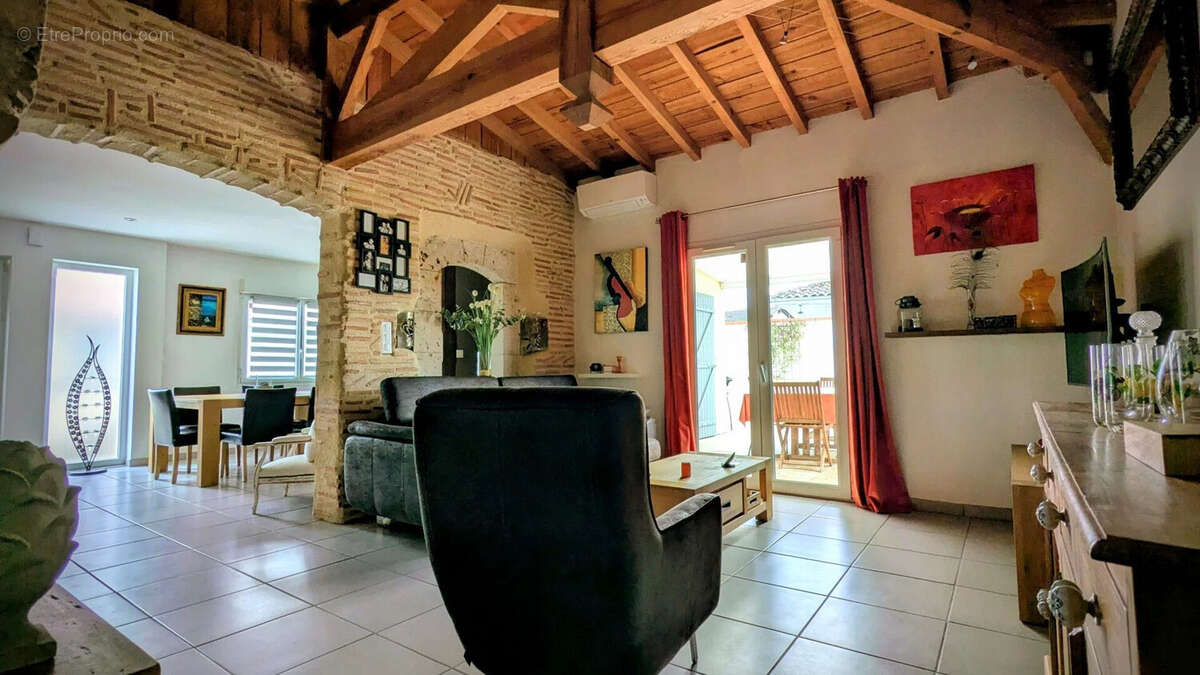 Un salon moderne avec des murs en briques, un plafond en bois et des meubles contemporains. Des oeuvres d'art décorent les murs, et une grande porte-fenêtre laisse entrer la lumière naturelle. - Maison à AIGUILLON