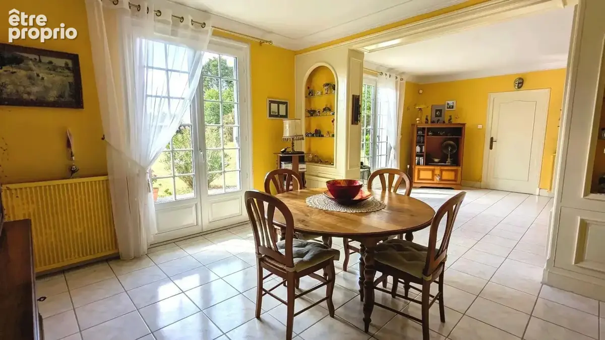 Salle à manger lumineuse avec des murs jaunes, une table ronde en bois et des chaises. Les fenêtres laissent entrer beaucoup de lumière naturelle.