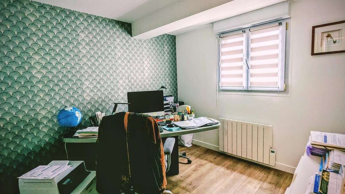 Un bureau avec un ordinateur, des papiers, et un globe sur le bureau. La pièce a un mur avec un motif géométrique et une fenêtre avec un store. - Maison à AIGUILLON