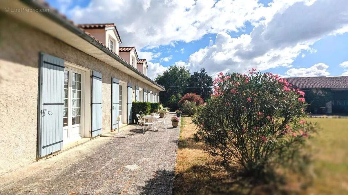 Une grande maison avec des volets bleus, une terrasse en pierre, des arbustes fleuris  - Maison à FOULAYRONNES