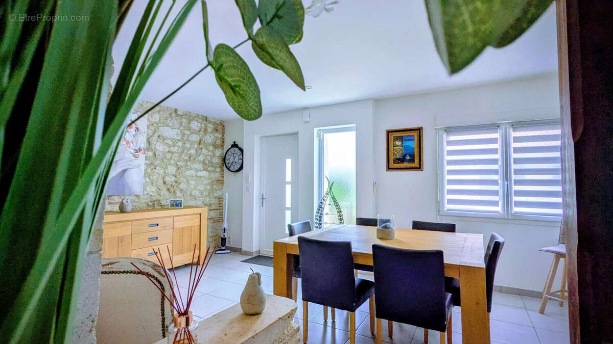 Salle à manger lumineuse avec table en bois, chaises noires, mur en pierre, et décorations végétales. - Maison à AIGUILLON