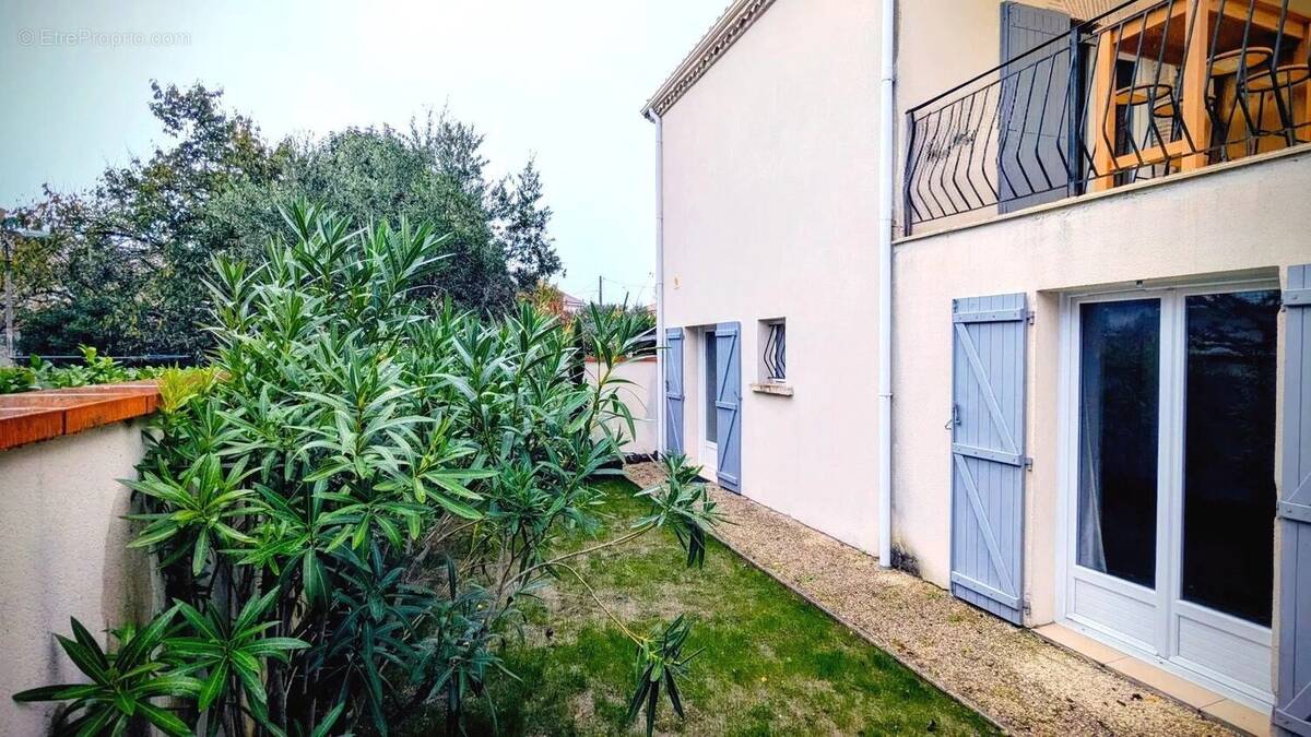 Il s'agit d'une maison avec un balcon et des volets bleus, entourée de verdure et située dans un jardin. - Maison à AIGUILLON