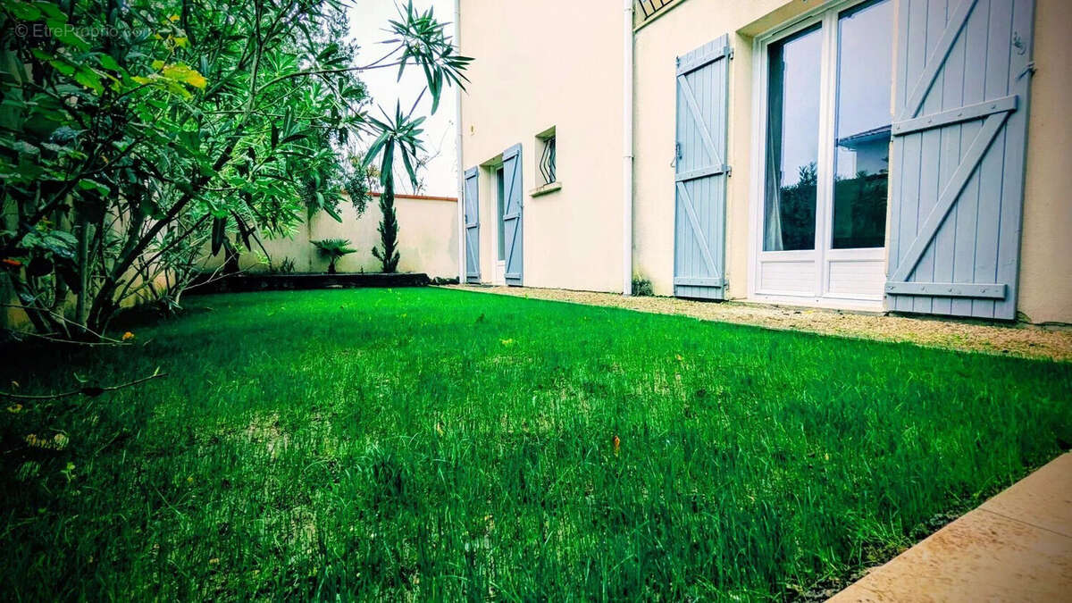 Jardin avec pelouse verte et arbustes près d'une maison aux volets bleus. - Maison à AIGUILLON