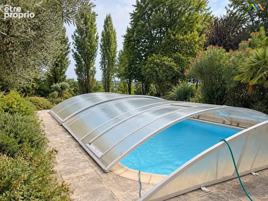 Une piscine sous abri bas, entourée d'arbres et de massifs.
