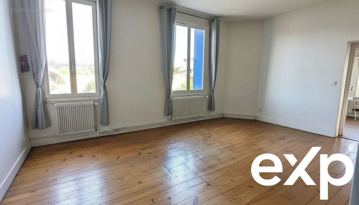 Une pièce lumineuse avec un sol en bois, deux grandes fenêtres avec rideaux gris et des murs blancs. - Maison à LE PASSAGE