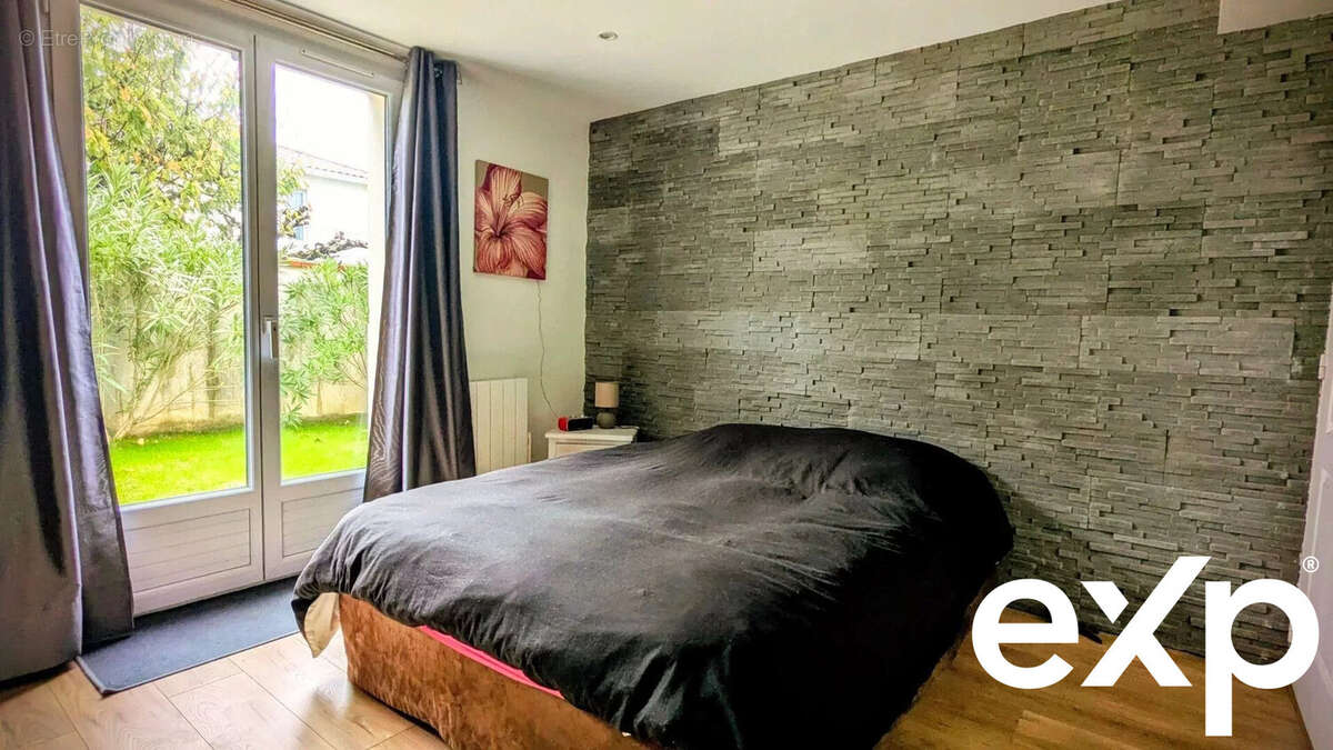 Chambre avec un mur en pierre, un lit noir, et une grande fenêtre donnant sur un jardin. - Maison à AIGUILLON