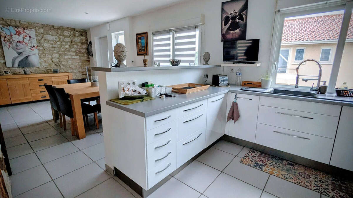 Cuisine moderne avec plan de travail blanc, salle à manger attenante et décorations murales. - Maison à AIGUILLON