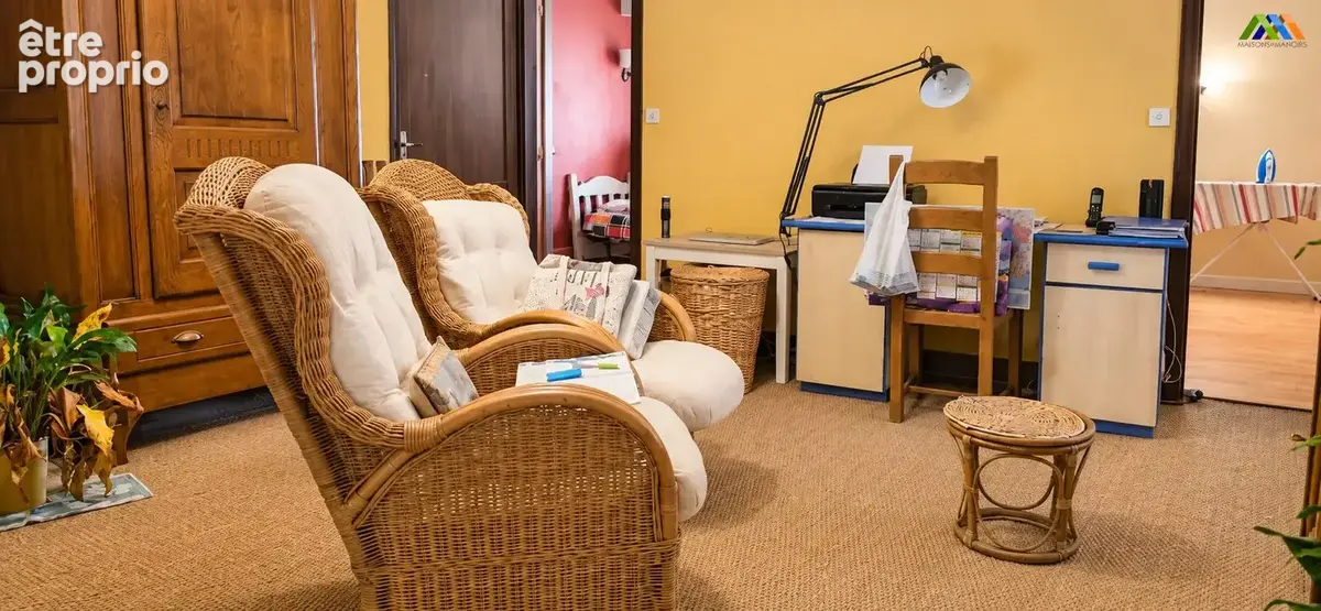 Coin salon aux murs jaunes, avec fauteuils en rotin, armoire en bois et espace bureau.