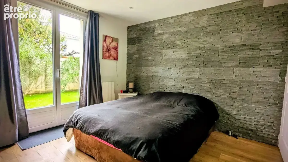 Chambre avec un mur en pierre, un lit noir, et une grande fenêtre donnant sur un jardin.