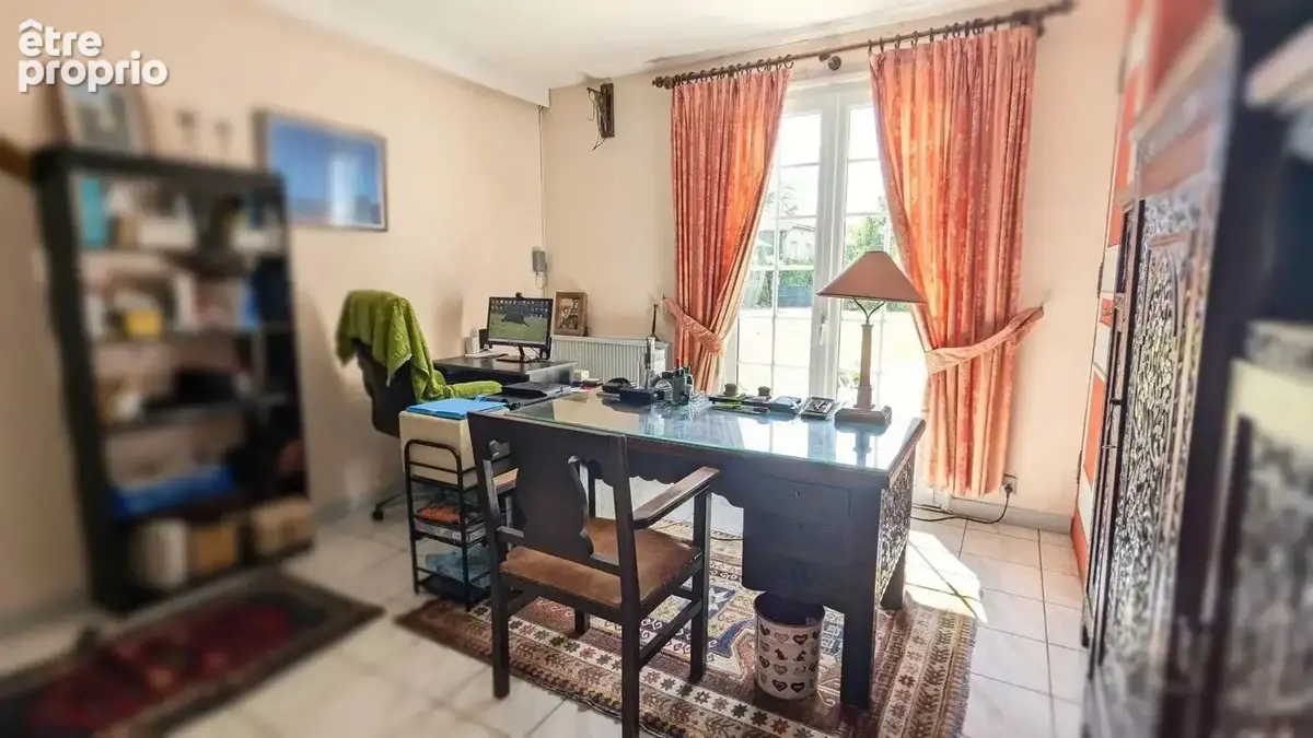 Un bureau lumineux avec un grand bureau en verre, des rideaux rouges, une étagère pleine de livres et une grande armoire sculptée à droite.