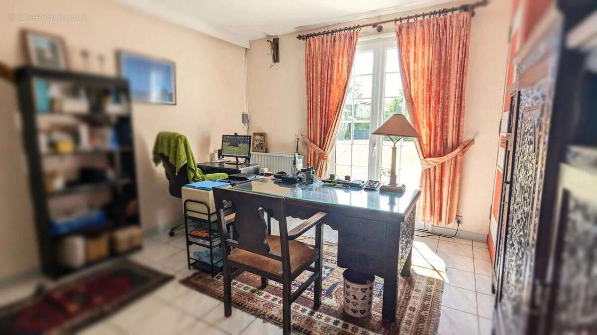Un bureau lumineux avec un grand bureau en verre, des rideaux rouges, une étagère pleine de livres et une grande armoire sculptée à droite. - Maison à FOULAYRONNES