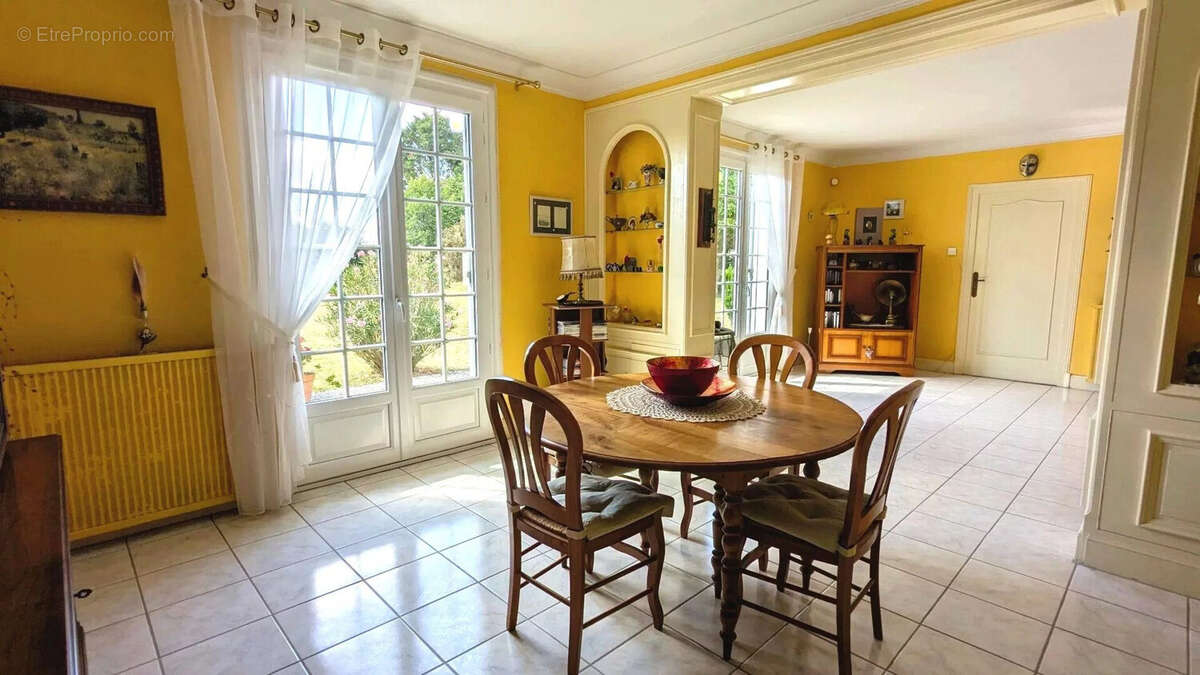 Salle à manger lumineuse avec des murs jaunes, une table ronde en bois et des chaises. Les fenêtres laissent entrer beaucoup de lumière naturelle. - Maison à FOULAYRONNES