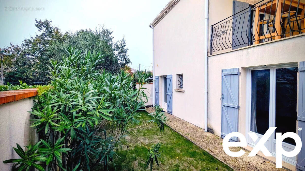 Il s'agit d'une maison avec un balcon et des volets bleus, entourée de verdure et située dans un jardin. - Maison à AIGUILLON