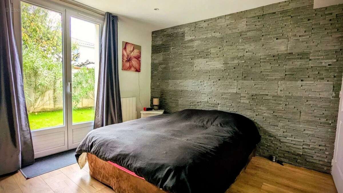 Chambre avec un mur en pierre, un lit noir, et une grande fenêtre donnant sur un jardin. - Maison à AIGUILLON