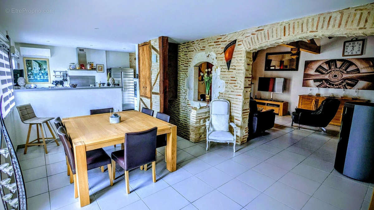 Une salle à manger ouverte avec une grande table en bois, des chaises noires, des murs en briques apparentes et une cuisine en arrière-plan. L'espace est moderne et lumineux. - Maison à AIGUILLON