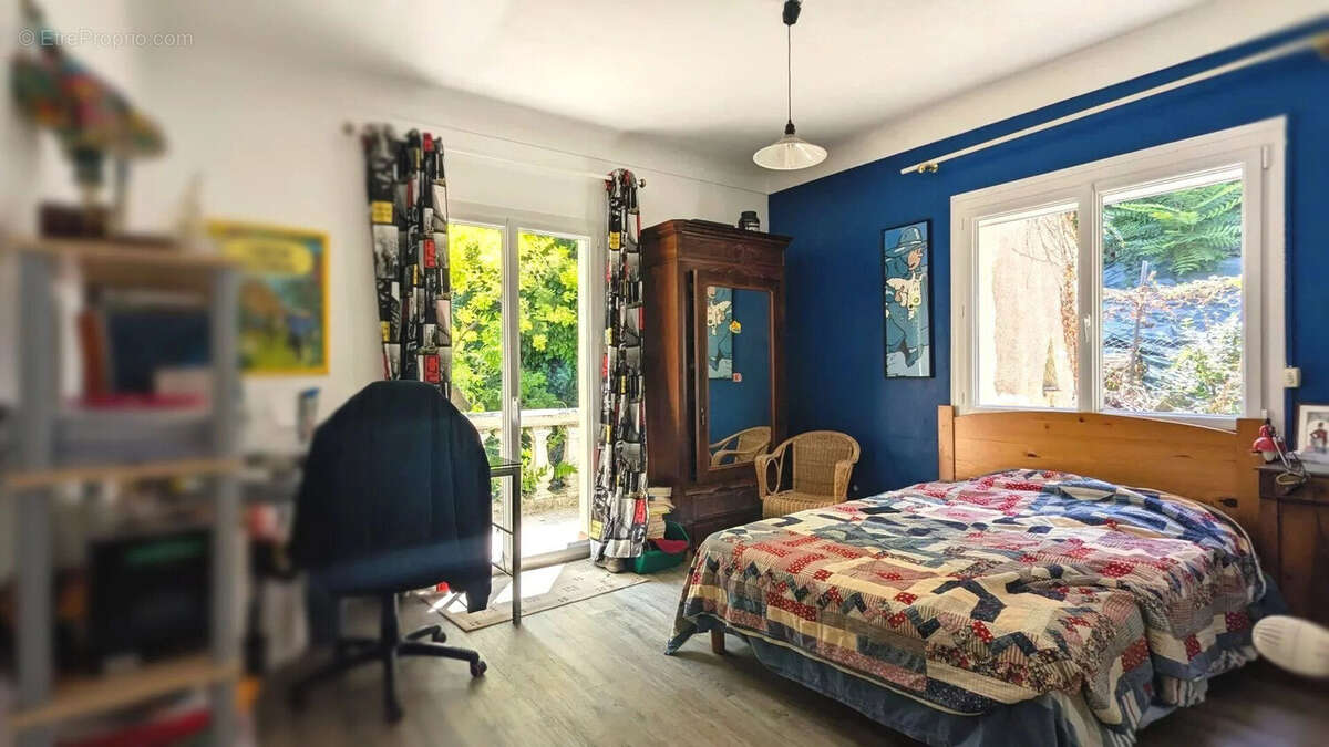 Chambre lumineuse avec un mur bleu, un lit coloré, une armoire en bois, et de grandes fenêtres donnant sur la verdure. - Maison à FOULAYRONNES