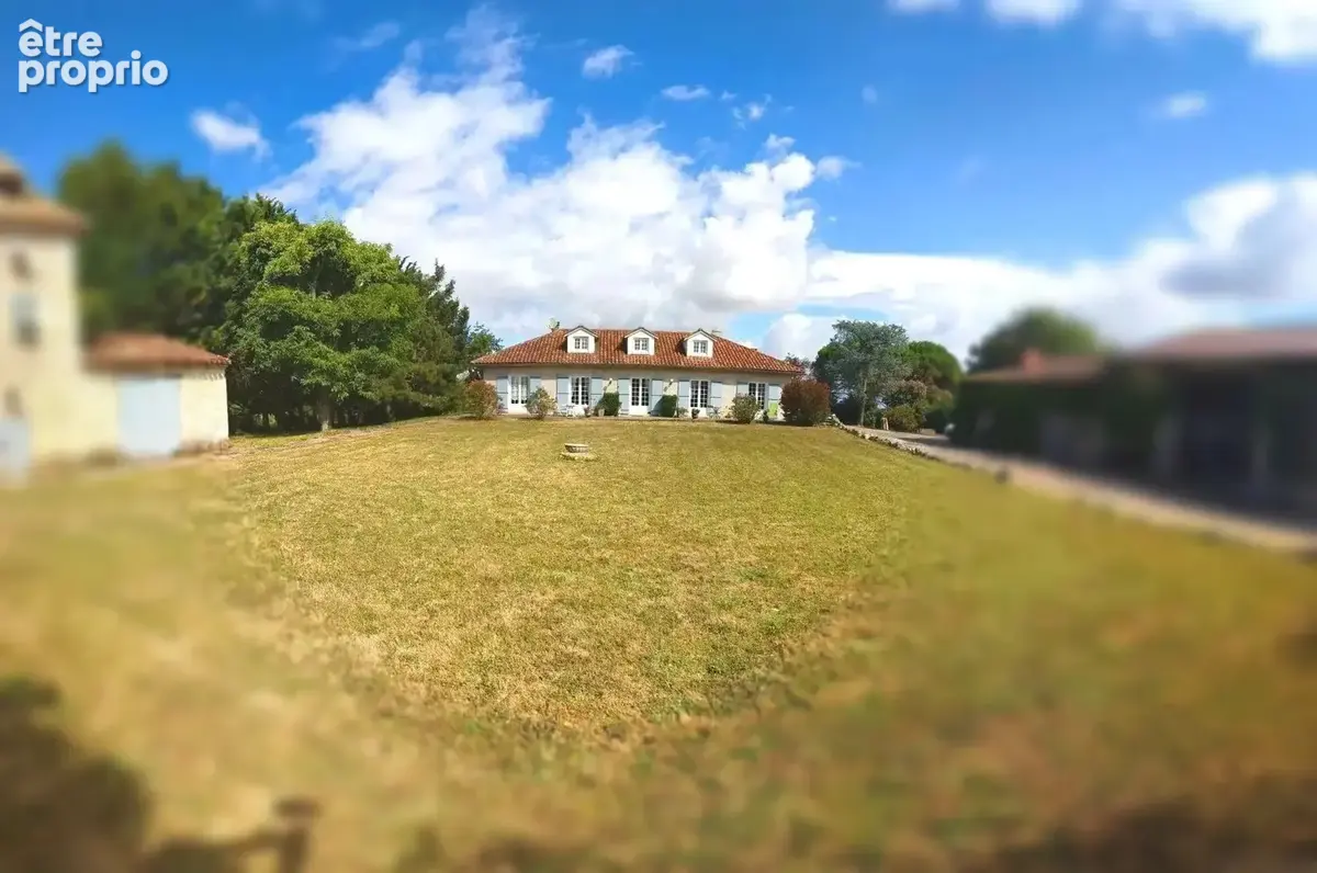 Une grande maison entourée de bâtiments en pierre, avec une pelouse verdoyante sous un ciel partiellement nuageux.