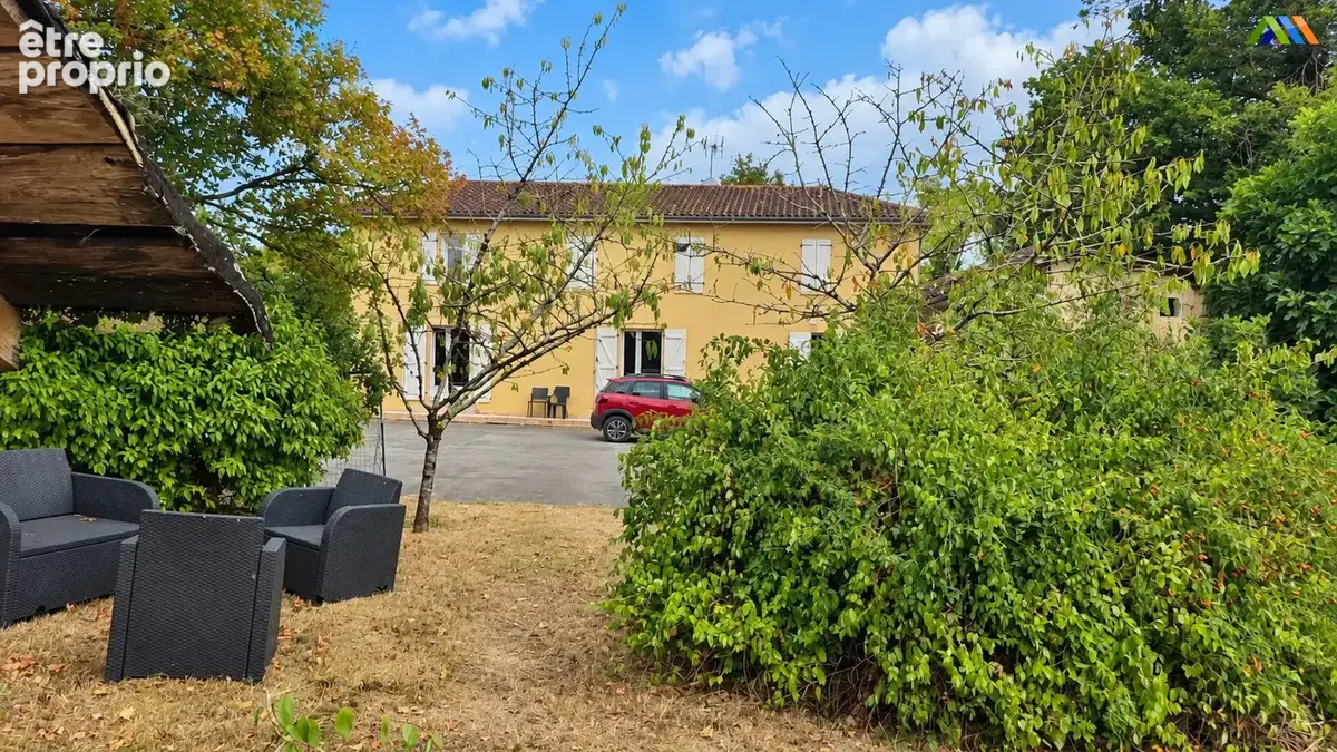 Jardin arboré avec coin détente extérieur, face à la façade jaune et aux volets blancs de la maison.