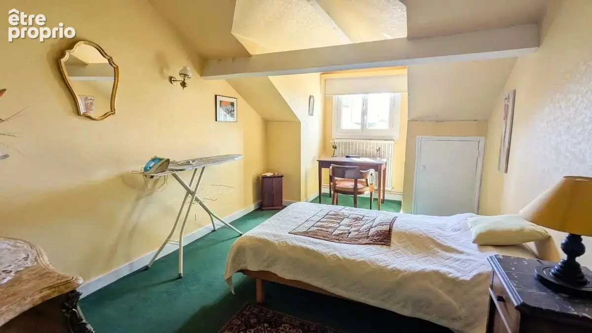 Une chambre lumineuse avec des murs jaunes, un lit simple, un fer à repasser sur sa table, et un bureau près d'une fenêtre.